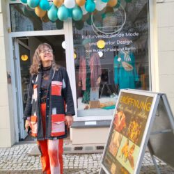 Frau in farbenfrohem Outfit mit rot-schwarz-weißen Akzenten steht vor einem Schaufenster mit Ballondekoration. Im Fenster steht: „Kulturverbindende Mode und Interior Design aus Burkina Faso und Berlin“. Rechts ein Aufsteller mit Plakat zur Eröffnung eines Ateliers.