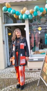 Frau in farbenfrohem Outfit mit rot-schwarz-weißen Akzenten steht vor einem Schaufenster mit Ballondekoration. Im Fenster steht: „Kulturverbindende Mode und Interior Design aus Burkina Faso und Berlin“. Rechts ein Aufsteller mit Plakat zur Eröffnung eines Ateliers.
