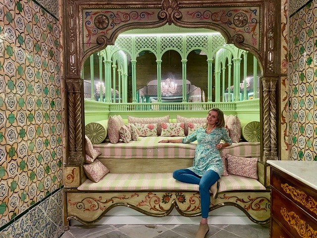 Eine Frau sitzt entspannt auf einer kunstvoll gestalteten, gepolsterten Bank in einer orientalisch dekorierten Nische mit floralen Mustern, geschnitztem Holz und grünen Arkaden im Hintergrund.