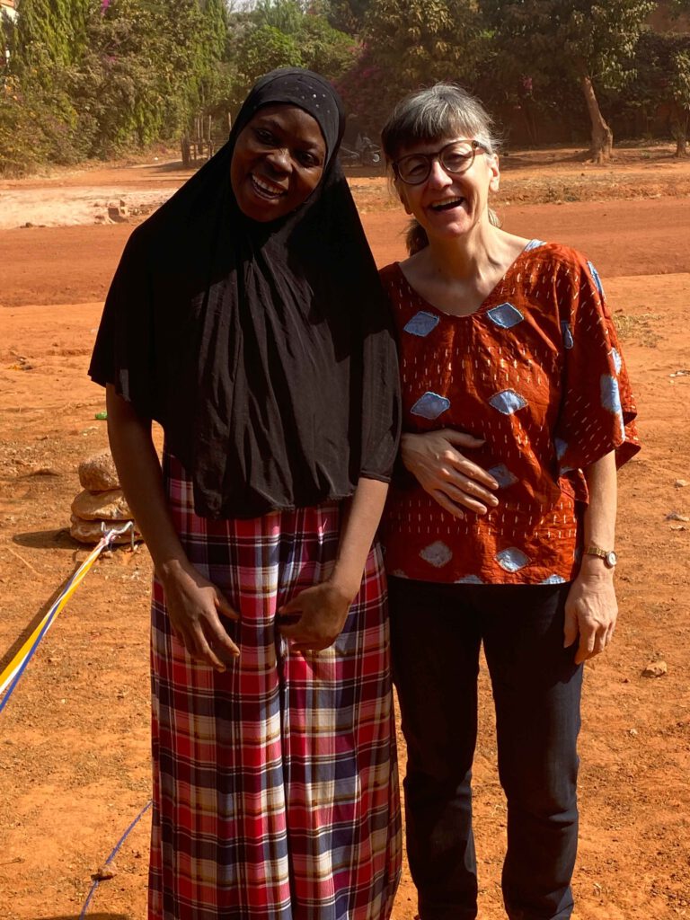 Zwei lachende Frauen, eine mit Kopftuch, stehen nebeneinander auf roter Erde in Burkina Faso, umgeben von Bäumen.
