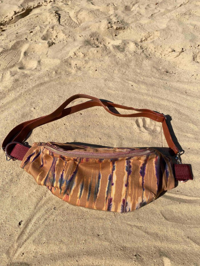 Bauchtasche aus afrikanischen Stoffen in Ocker- und Violetttönen mit braunem Riemen, liegend auf hellem Sand.