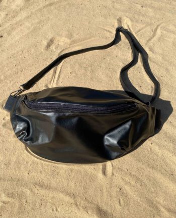 Schwarze, handgefertigte Bauchtasche aus glattem Kunstleder mit verstellbarem Riemen, liegend auf hellem Sand.
