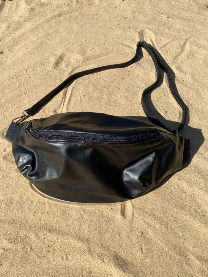 Schwarze, handgefertigte Bauchtasche aus glattem Kunstleder mit verstellbarem Riemen, liegend auf hellem Sand.