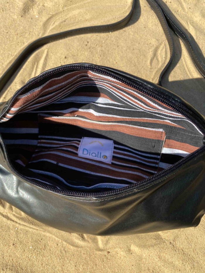Offene Bauchtasche mit schwarzer Außenseite und Innenfutter aus gestreiftem Baumwollgewebe "Faso dan Fani" in Braun-, Schwarz- und Weiß, mit Label „Diallo tissue tales“, liegend auf Sand.