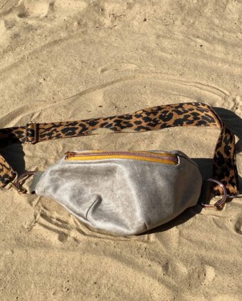 Elegante graue Bauchtasche aus Leder mit gelbem Reißverschluss und breitem, gemustertem Gurt in Leopardenoptik, liegend auf hellem Sand.