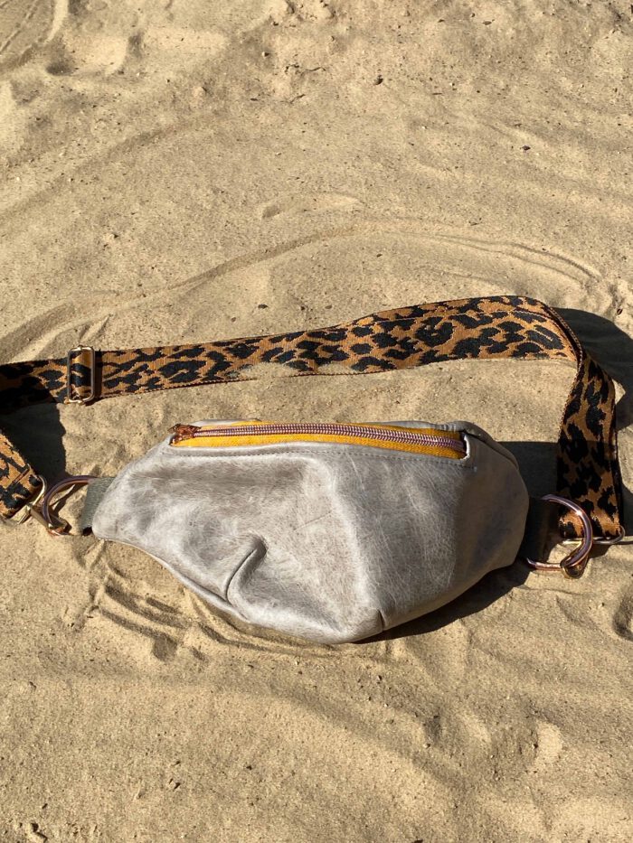 Elegante graue Bauchtasche aus Leder mit gelbem Reißverschluss und breitem, gemustertem Gurt in Leopardenoptik, liegend auf hellem Sand.