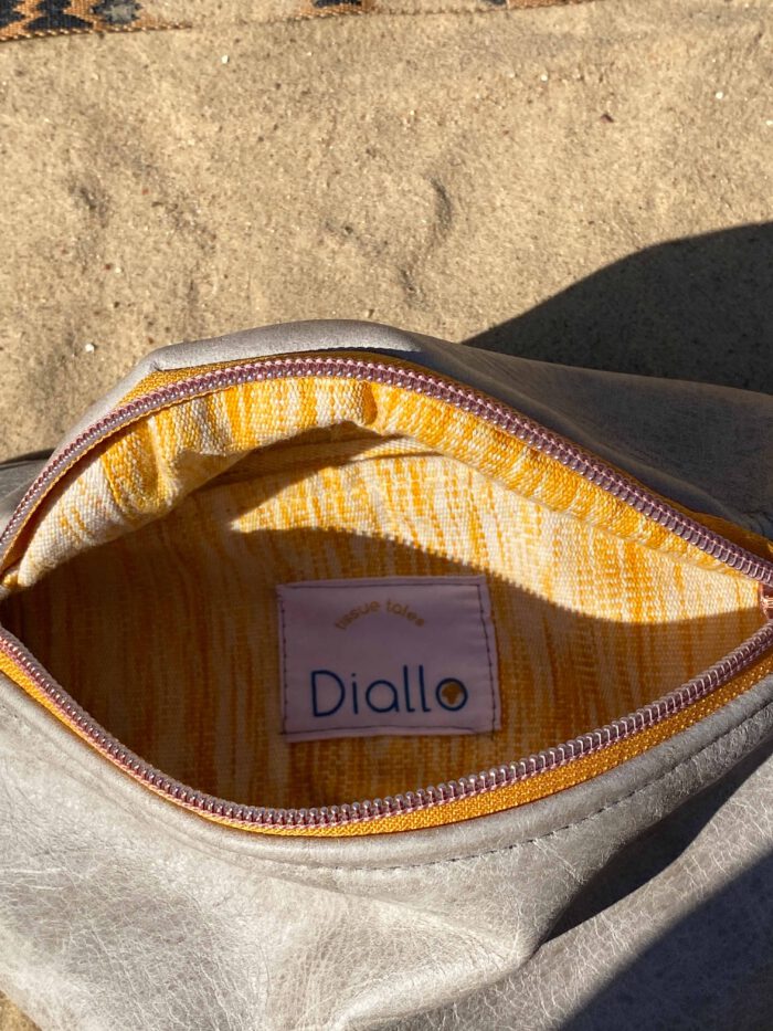 Geöffnete Bauchtasche aus grauem Leder mit gelbem Innenfutter aus handgewebtem afrikanischen Stoff und Label „Diallo tissue tales“, liegend auf Sand.