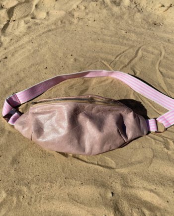 Rosafarbene Lederbauchtasche mit verstellbarem rosa-weiß gestreiftem Gurt, liegend auf hellem Sand.