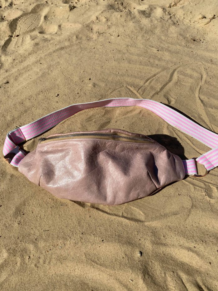 Rosafarbene Lederbauchtasche mit verstellbarem rosa-weiß gestreiftem Gurt, liegend auf hellem Sand.