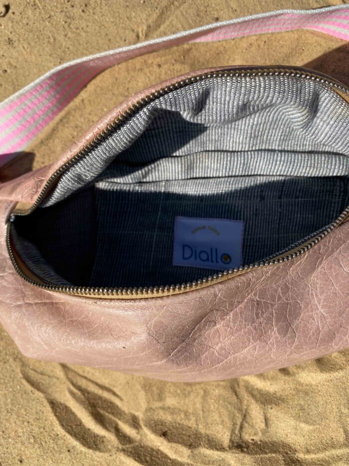 Geöffnete rosafarbene Lederbauchtasche mit grauem Innenfutter aus handgewebtem Faso dan Fani und Label „Diallo tissue tales“, liegend auf Sand.