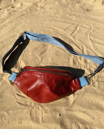 Rote Leder-Bauchtasche (Belly Bag) mit hellblauem Riemen liegt auf hellem Sand, inspiriert von afrikanischem Batikstoff.