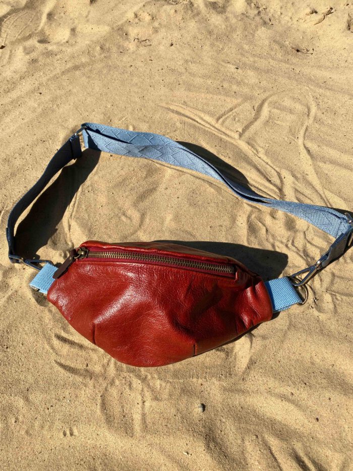 Rote Leder-Bauchtasche (Belly Bag) mit hellblauem Riemen liegt auf hellem Sand, inspiriert von afrikanischem Batikstoff.