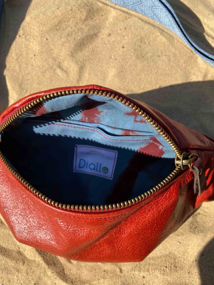 Offene rote Leder-Bauchtasche (Belly Bag) mit hellblauem Riemen liegt auf hellem Sand, das Innenfutter ist aus afrikanischem Batikstoff in rot und blau.