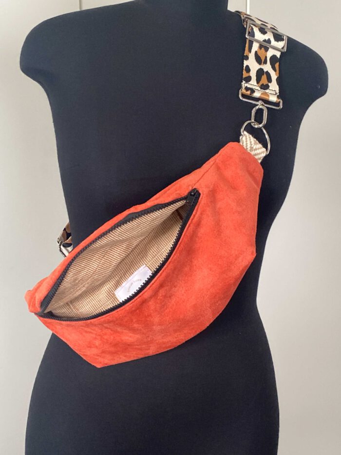 Wildleder-Bauchtasche (Belly Bag) in Flamingoton mit schwarzem Reißverschluss, geöffnet und mit beige-gestreiftem Innenfutter, sowie einem breiten Leopardenriemen, inspiriert von afrikanischer Mode, präsentiert auf einer schwarzen Schneiderpuppe.