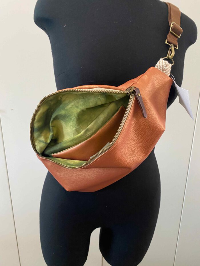 Kupferfarbene Kunstleder-Bauchtasche (Belly Bag) mit geöffnetem goldfarbenem Reißverschluss, grün gemustertem Innenfutter und braunem Schulterriemen, inspiriert von afrikanischer Mode, präsentiert auf einer schwarzen Schneiderpuppe.