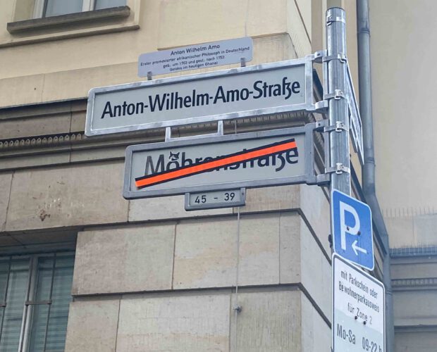 Straßenschild mit der Aufschrift „Anton-Wilhelm-Amo-Straße“ in Berlin, darüber ein Zusatzschild mit Informationen über Anton Wilhelm Amo, den ersten afrikanischen promovierten Philosophen in Deutschland. Das durchgestrichene Schild „Mohrenstraße“ ist noch sichtbar.