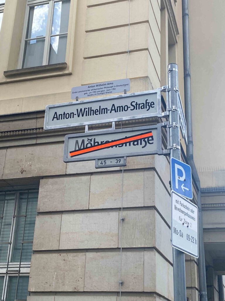 Straßenschild mit der Aufschrift „Anton-Wilhelm-Amo-Straße“ in Berlin, darüber ein Zusatzschild mit Informationen über Anton Wilhelm Amo, den ersten afrikanischen promovierten Philosophen in Deutschland. Das durchgestrichene Schild „Mohrenstraße“ ist noch sichtbar.