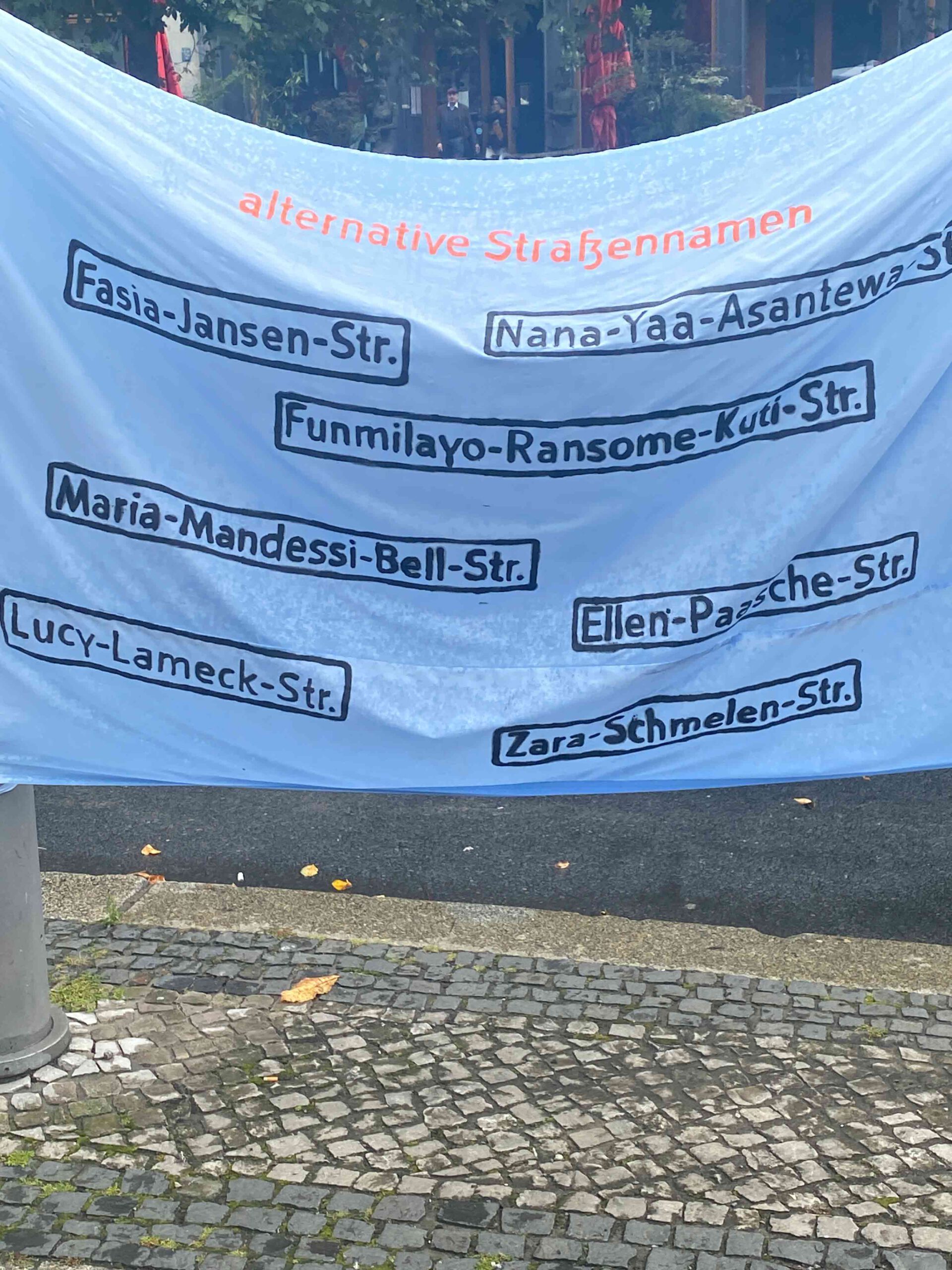 Transparent mit der Aufschrift „alternative Straßennamen“, darauf mehrere Vorschläge wie „Fasia-Jansen-Str.“, „Nana-Yaa-Asantewaa-Str.“, „Funmilayo-Ransome-Kuti-Str.“ und weitere.