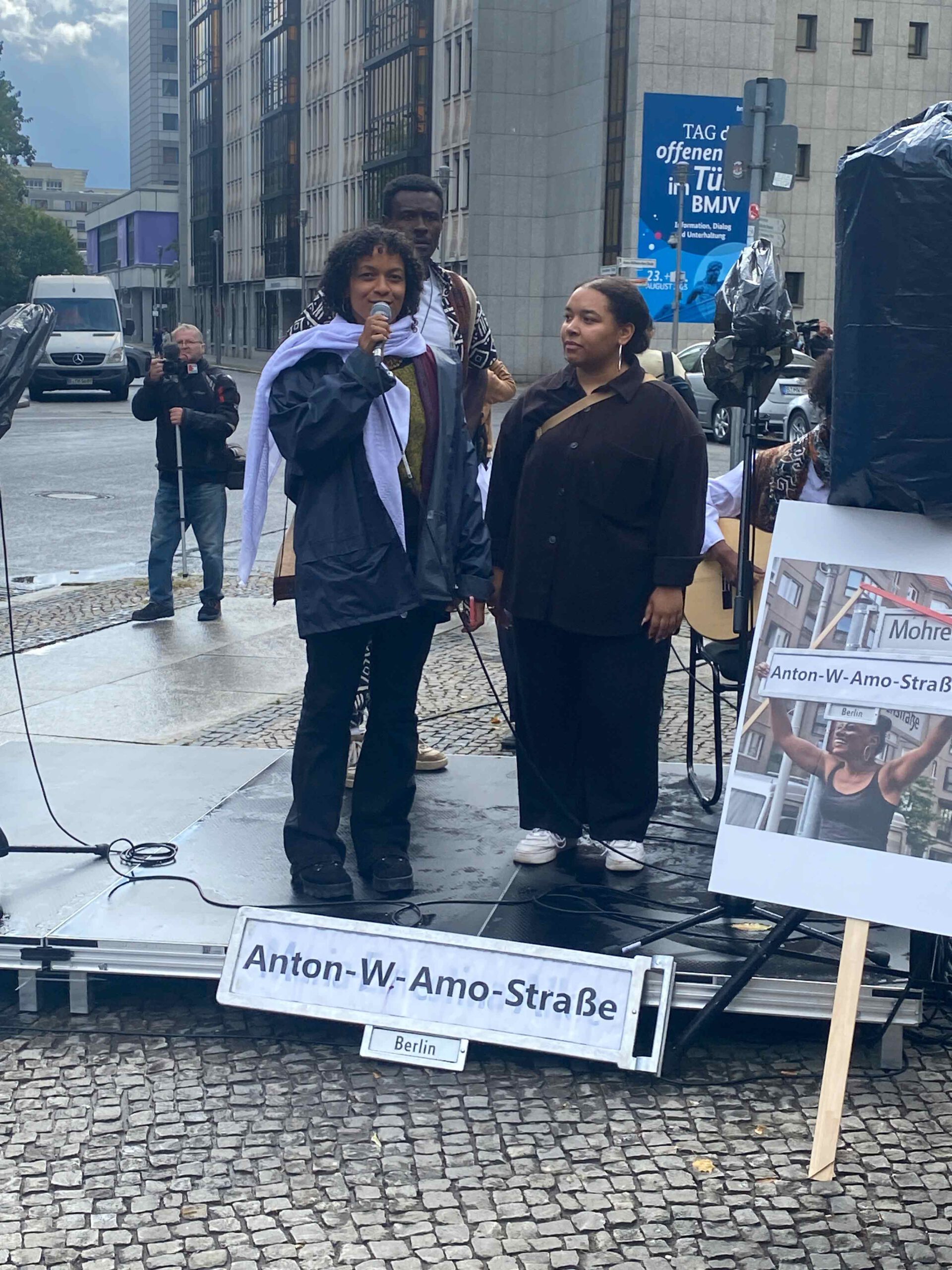 Menschen sprechen bei einer Kundgebung mit Mikrofon, im Vordergrund ein Schild mit „Anton-W.-Amo-Straße“. Im Hintergrund stehen weitere Personen sowie Transparente.