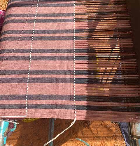 Handgewebter Stoff in Rosé- und Schwarztönen auf einem traditionellen Webstuhl. Die Streifen verlaufen längs und quer, wodurch ein dezentes Karomuster entsteht. Im Vordergrund ist der Webprozess zu sehen, bei dem der Stoff Stück für Stück entsteht, während Sonnenlicht die Farben warm hervorhebt.