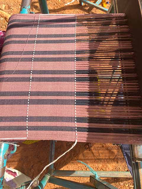 Handgewebter Stoff in Rosé- und Schwarztönen auf einem traditionellen Webstuhl. Die Streifen verlaufen längs und quer, wodurch ein dezentes Karomuster entsteht. Im Vordergrund ist der Webprozess zu sehen, bei dem der Stoff Stück für Stück entsteht, während Sonnenlicht die Farben warm hervorhebt.
