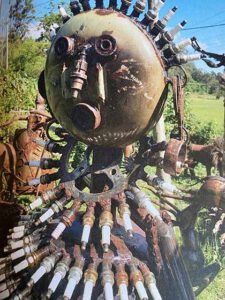 Skulptur aus alten Metallteilen und Zündkerzen, die eine menschliche Figur mit rundem Kopf darstellt, im Freien aufgestellt.