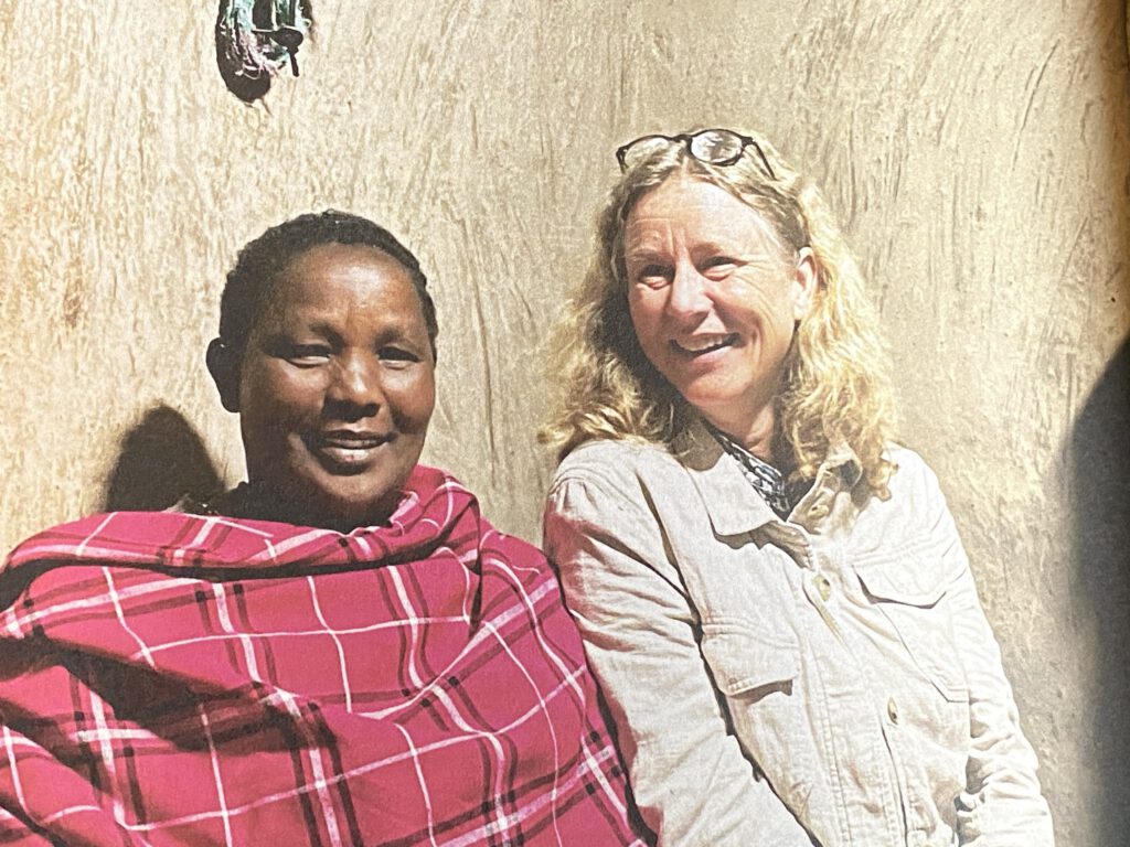 Zwei Frauen sitzen nebeneinander und lächeln: eine Frau in rotem kariertem Tuch, die andere mit blonden Haaren und heller Jacke. Sie sitzen vor einer Wand aus Lehm oder Ton.