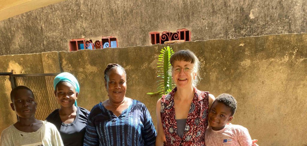 Sechs Menschen unterschiedlichen Alters stehen nebeneinander auf einer gefliesten Terrasse. Vier Kinder und zwei erwachsene Frauen lächeln in die Kamera. Sie tragen farbenfrohe, teilweise gemusterte Kleidung. Im Hintergrund ist eine Hauswand mit kleinen Fenstern und Pflanzen zu sehen, das Licht ist warmes Tageslicht.