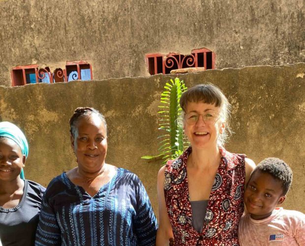 Sechs Menschen unterschiedlichen Alters stehen nebeneinander auf einer gefliesten Terrasse. Vier Kinder und zwei erwachsene Frauen lächeln in die Kamera. Sie tragen farbenfrohe, teilweise gemusterte Kleidung. Im Hintergrund ist eine Hauswand mit kleinen Fenstern und Pflanzen zu sehen, das Licht ist warmes Tageslicht.