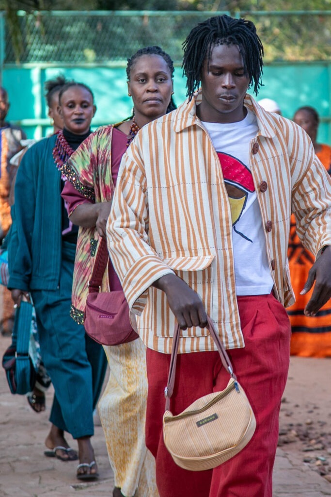 Mehrere Personen gehen in einer Reihe über einen Hof. Im Vordergrund eine Person mit gestreifter Jacke, roter Hose und kleiner Tasche, dahinter weitere Personen in farbenfroher Kleidung.