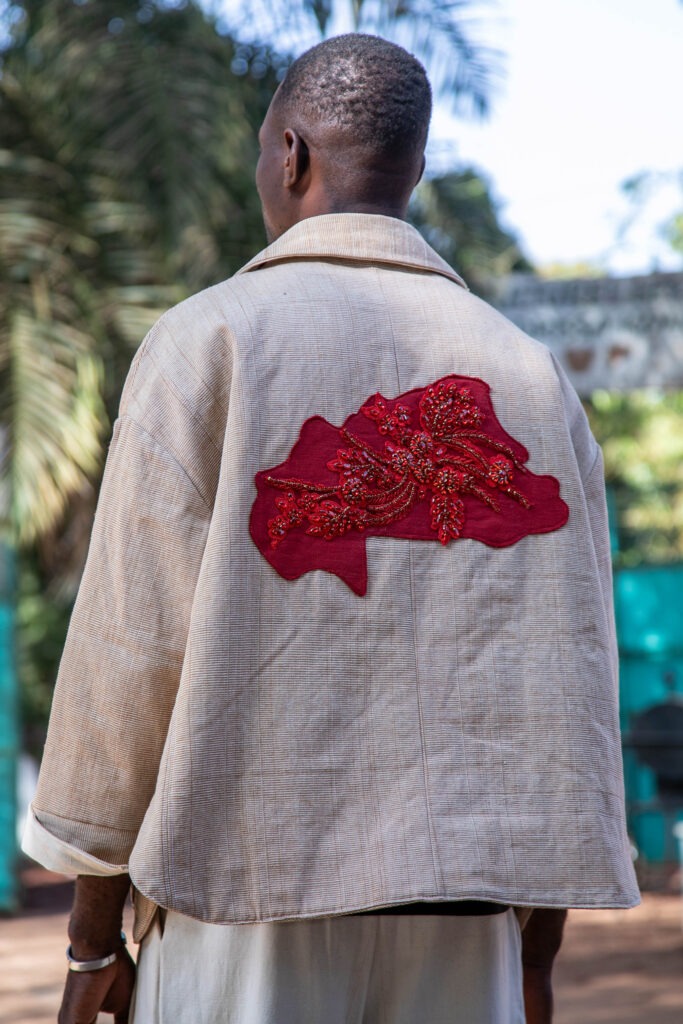 Person von hinten in beigefarbener Jacke mit aufwendig besticktem rotem Stoffelement auf dem Rücken, im Freien vor tropischer Vegetation.