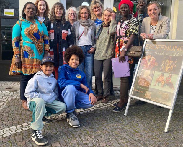 Gruppenfoto vor einem festlich geschmückten Ladeneingang mit Ballongirlande: Zehn Erwachsene und zwei Kinder posieren vor einem Aufsteller zur Eröffnung von „Diallo tissue tales – Shop, Events & Atelier“. Die Gruppe lächelt in die Kamera, einige tragen farbenfrohe Kleidung; die Szene wirkt einladend, feierlich und gemeinschaftlich.