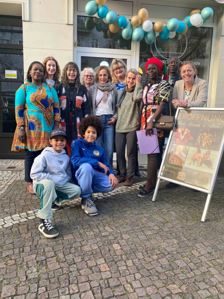 Gruppenfoto vor einem festlich geschmückten Ladeneingang mit Ballongirlande: Zehn Erwachsene und zwei Kinder posieren vor einem Aufsteller zur Eröffnung von „Diallo tissue tales – Shop, Events & Atelier“. Die Gruppe lächelt in die Kamera, einige tragen farbenfrohe Kleidung; die Szene wirkt einladend, feierlich und gemeinschaftlich.