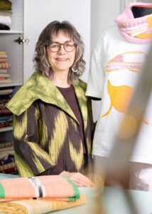 Alternativtext: **Porträt einer lächelnden Frau mit Brille in einem Atelier oder Stoffladen. Sie trägt ein gemustertes grün-braunes Kleidungsstück und steht neben einer Schneiderpuppe mit weißem Oberteil und rosa Schal; im Hintergrund sind gefaltete Stoffe zu sehen.**