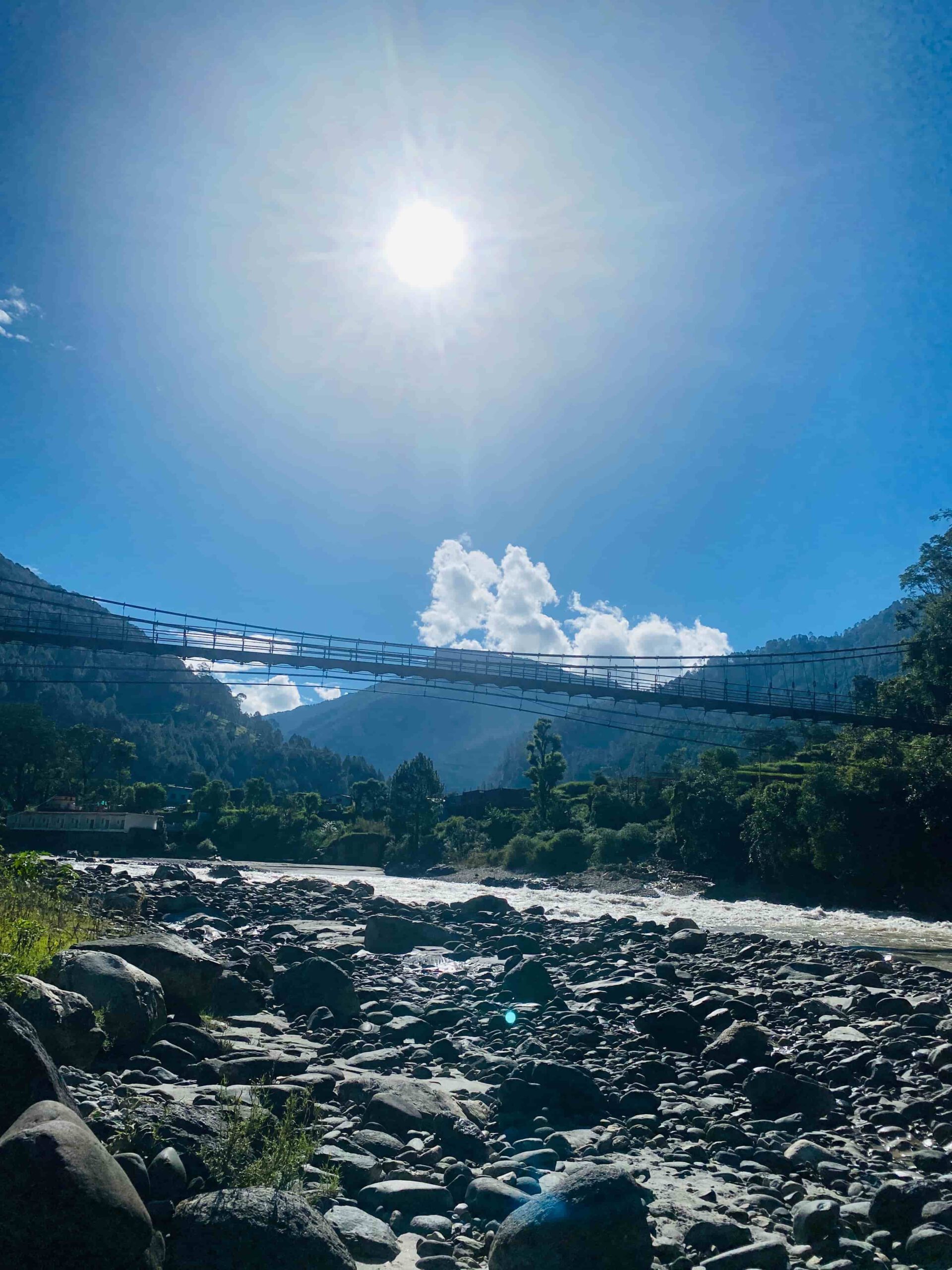 Eine Hängebrücke spannt sich über das Flussbett des Ganges, dahinter strahlender Sonnenschein und ein imposantes Wolkenkonstrukt. das Motiv symbolisiert den Aufbruch zu einer Reise zu sich selbst.